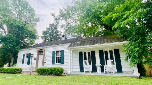 A charming white house with green shutters and rocking chairs on the porch surrounded by greenery.