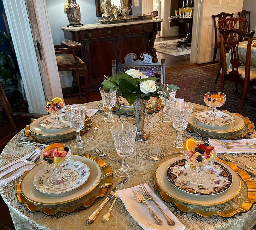 An elegantly set dining table with floral centerpieces, dessert cups, and fine china in a well-decorated room.
