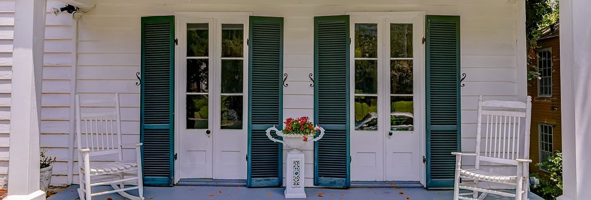 A porch featuring two rocking chairs, double doors with green shutters, and a flower pot.