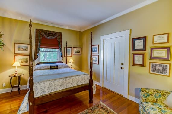 A cozy bedroom with a four-poster bed, warm yellow walls, and framed artwork.